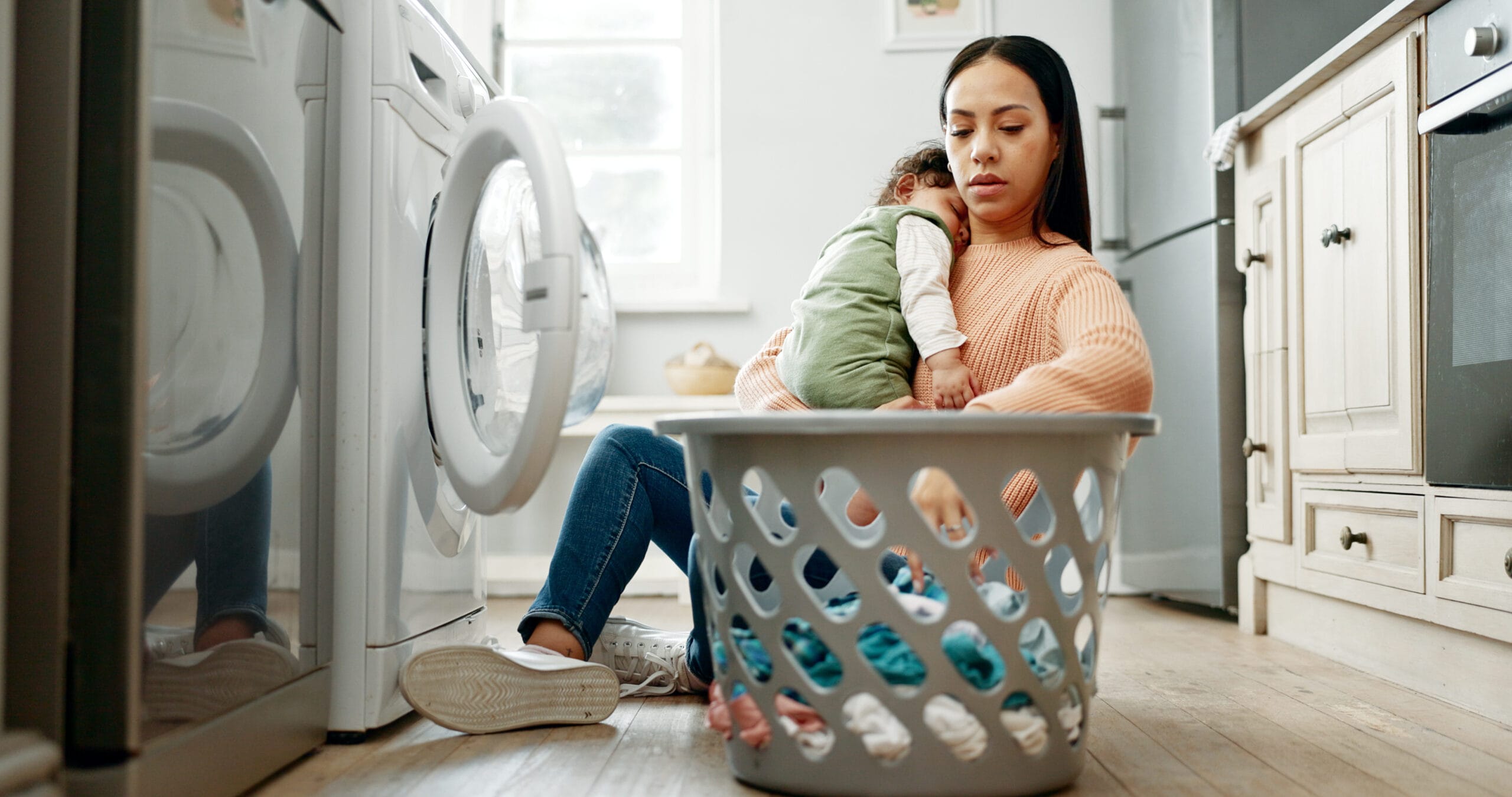 Mother, baby and basket in kitchen for laundry, housekeeping and motherhood in house with sleeping child. Woman, newborn and tired with washing machine for cleaning clothes, parenthood and burnout birth trauma